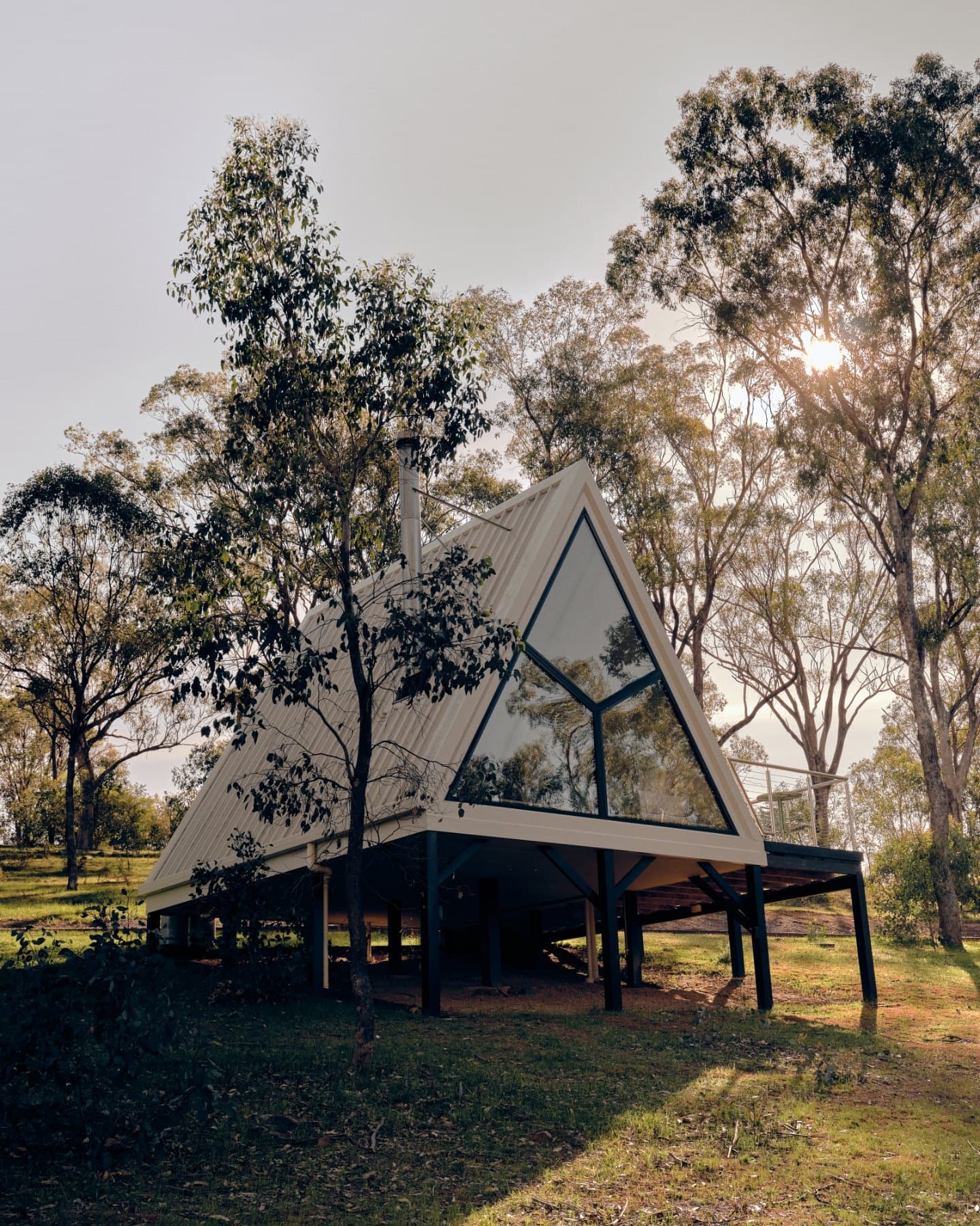 An A-Frame cabin on its piers among the gum trees, the sun low behind it