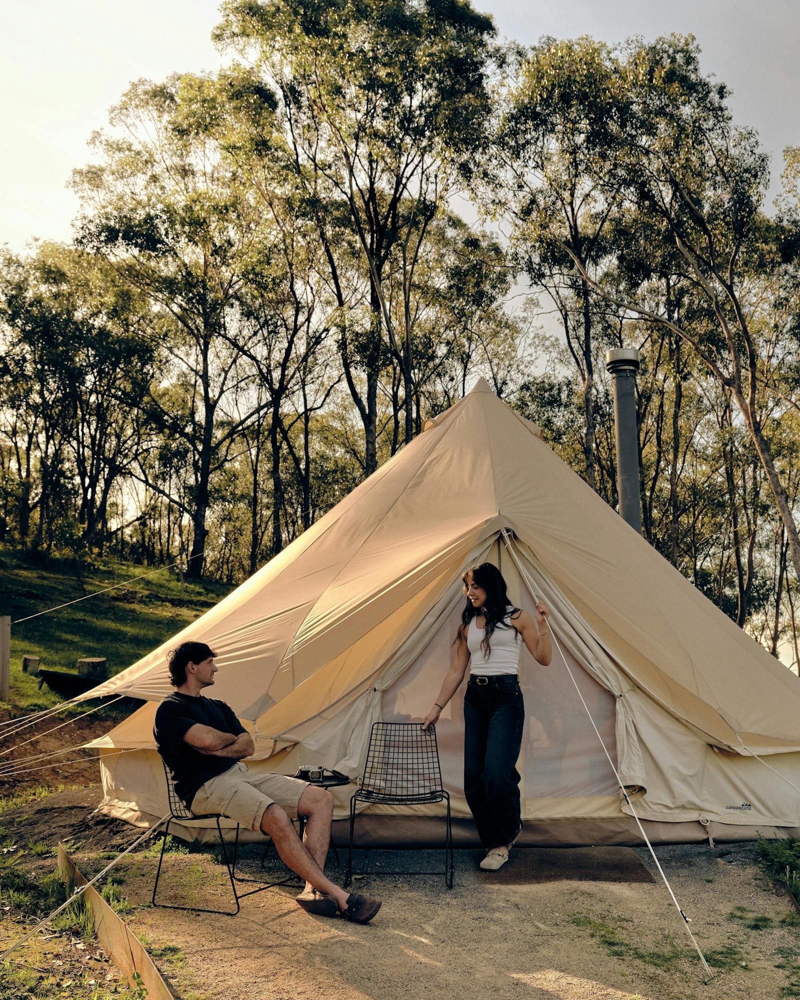 A couple at the door of a Luxe bell tent, the wood heater's flue rising beside the canvas
